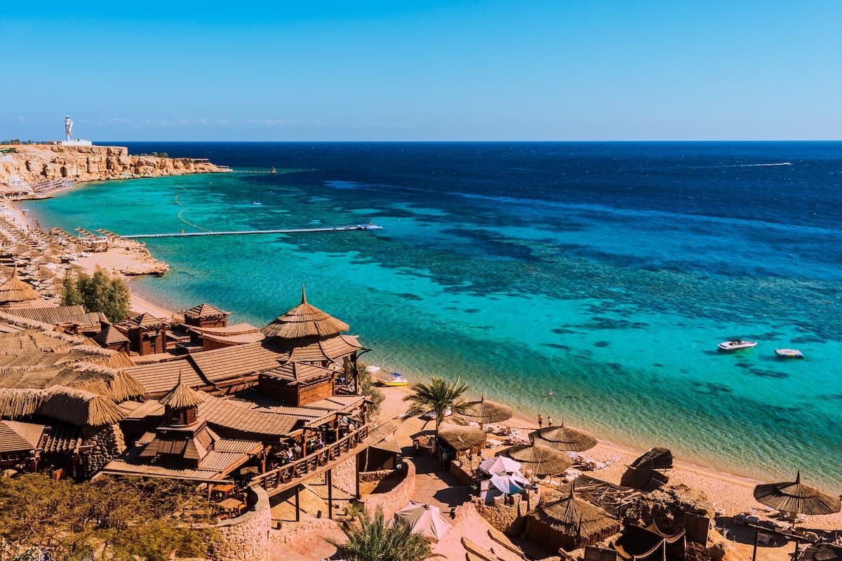 Flyfoto av en bukt ved Sharm el-Sheikh ved Rødehavet — restauranter på pæler, turkist vann med korallrev, brygger.